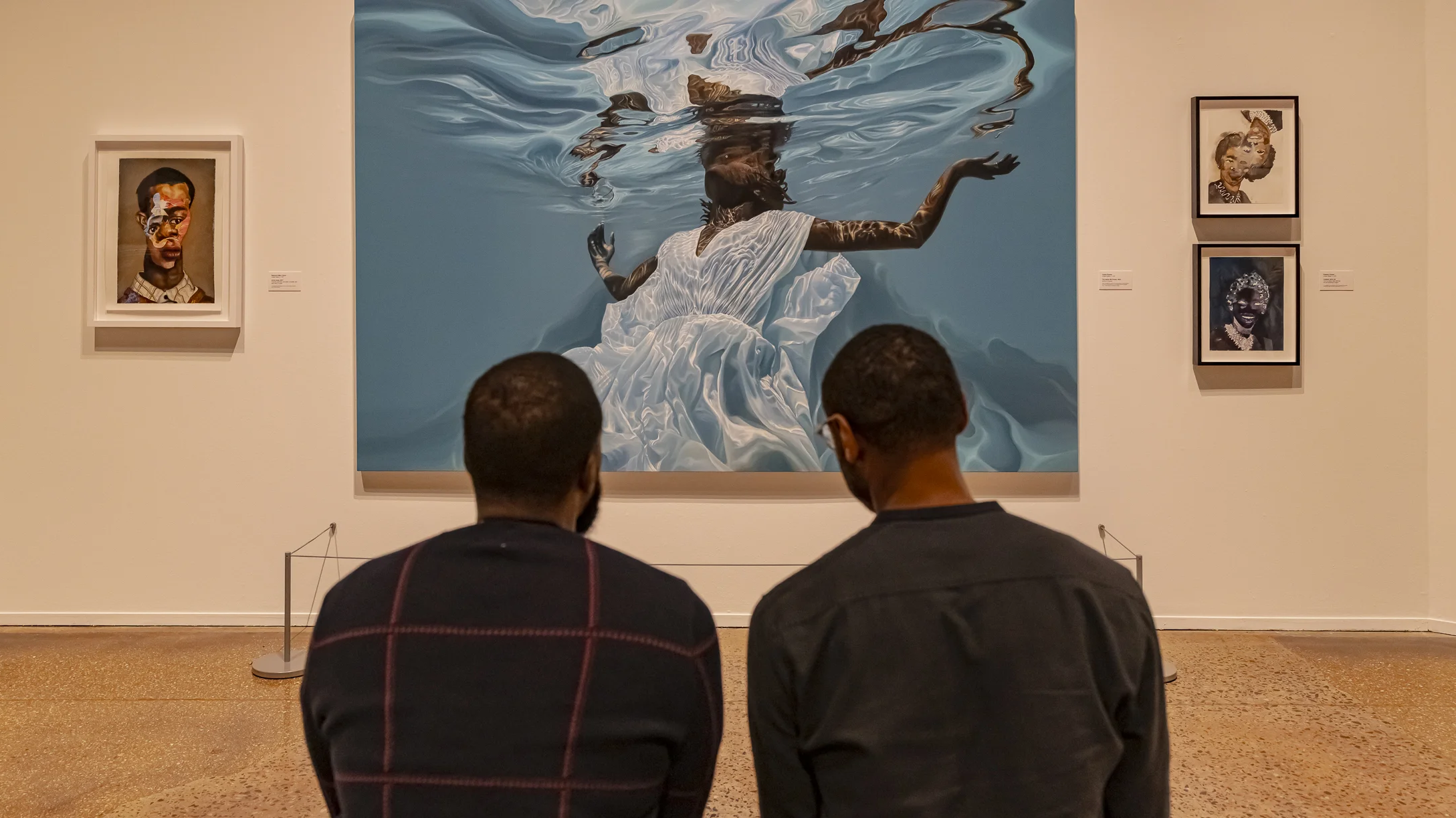 Two men sitting in gallery observing a large painting of a woman floating in water